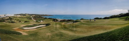 Foto de Sotogrande, España - 23 de enero de 2024 - Una vista panorámica de un campo de golf con golfistas, trampas de arena y una vista clara del mar y la costa bajo un cielo azul. - Imagen libre de derechos