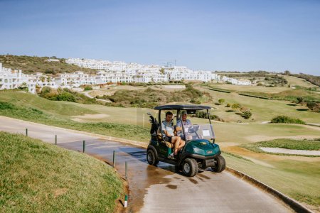 Téléchargez les photos : Sotogrande, Espagne - 23 janvier 2024 - Deux golfeurs dans une voiturette de golf sur un sentier avec un vaste terrain de golf et des bâtiments résidentiels blancs en arrière-plan. - en image libre de droit