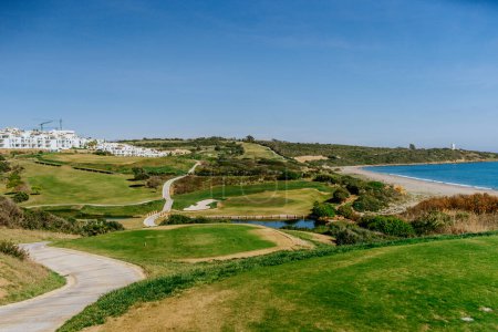 Foto de Sotogrande, España - 23 de enero de 2024 - Un campo de golf con un sendero curvo conduce hacia el mar, con una playa a la derecha y edificios residenciales a la izquierda bajo un cielo azul. - Imagen libre de derechos