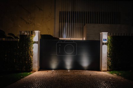 Sotogrande, Spanien - 23. Januar 2024 - Nachtansicht des Eingangs eines Luxus-Hauses mit schwarzem Schiebetor, weißen Wänden und weicher Beleuchtung.