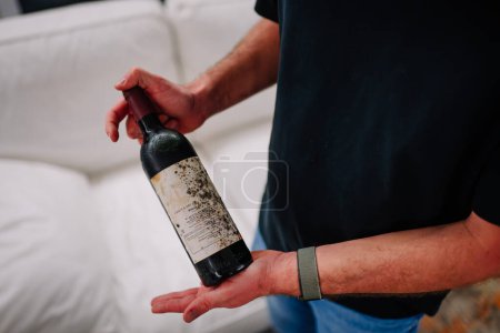 Photo for Sotogrande, Spain - January, 23, 2024 - A person is holding a dusty wine bottle with a label, standing against a blurred background with a white couch. - Royalty Free Image
