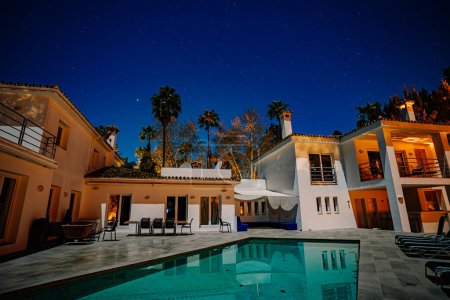 Téléchargez les photos : Villa de style méditerranéen avec piscine la nuit, étoiles, intérieur éclairé, meubles de patio, palmiers, ciel clair. - en image libre de droit