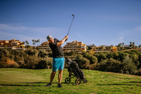 Photo for Sotogrante, Spain - January 25, 2024 - Man in mid-swing at golf, ball in air, pushcart beside, buildings in background, clear sky. - Royalty Free Image