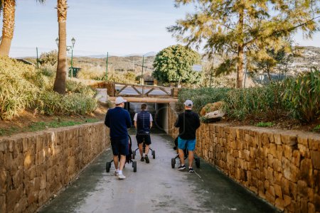 Téléchargez les photos : Sotogrante, Espagne - 25 janvier 2024 - Trois personnes marchent sur un sentier, poussant des sacs de golf, entourés de murs de pierre et de plantes. - en image libre de droit