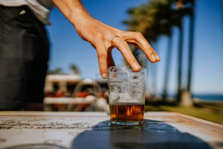 Foto de Sotogrante, España - 25 de enero de 2024 - Una mano que toma un vaso medio lleno de cerveza sobre una mesa con palmeras y un cielo azul al fondo. - Imagen libre de derechos