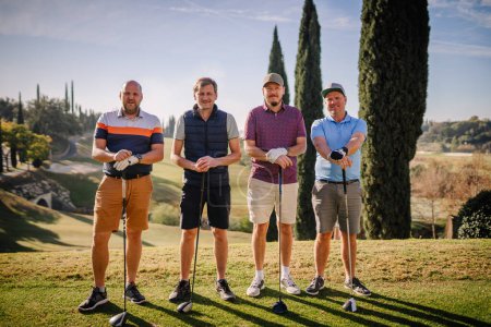 Foto de Sotogrante, España - 26 de enero de 2024 - cuatro golfistas con palos, alineados y sonriendo en un campo de golf, con cipreses y un cielo despejado en el fondo. - Imagen libre de derechos