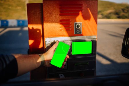 Photo for Sotogrante, Spain - January 26, 2024 -  hand reaching out from a car window holding a smartphone with a green screen towards a toll booth machine. - Royalty Free Image