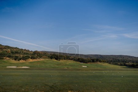 Sotogrante, Spain - January 27, 2024 - A golf course driving range with a green, scattered white golf balls, sand bunkers, trees, and hills under a vast blue sky.