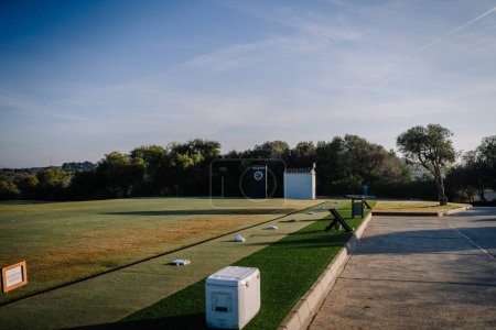 Sotogrante, Spain - January 27, 2024 -A driving range with scattered golf balls on artificial turf, a cooler, and a distance marker sign under a clear sky.