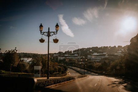 Sotogrante, Spain - January 27, 2024 - Ornate street lamps with hanging baskets on a road curving into a residential area, with a sun flare and scattered clouds in the sky.