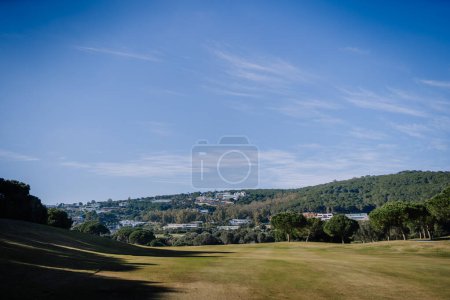 Sotogrante, Spain - January 27, 2024 - Golf course stretching towards a tree-covered hill with buildings on top under a spacious blue sky.
