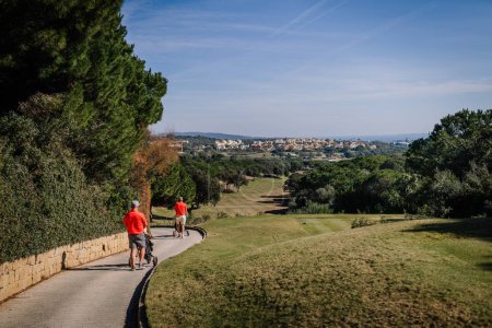 Téléchargez les photos : Sotogrante, Espagne - 27 janvier 2024 - Deux golfeurs en chemises rouges marchant sur un sentier avec des sacs de golf, surplombant un terrain de golf et un paysage urbain lointain - en image libre de droit