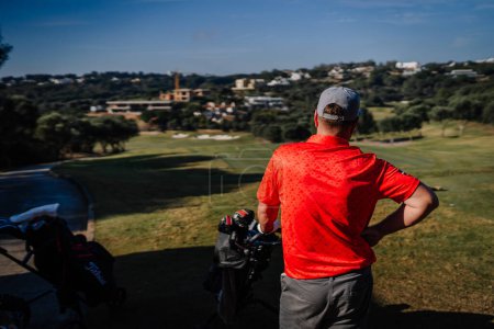 Téléchargez les photos : Sotogrante, Espagne - 27 janvier 2024 - Homme en chemise rouge et casquette observant un parcours de golf, des sacs de golf au premier plan et des collines avec des maisons au loin. - en image libre de droit