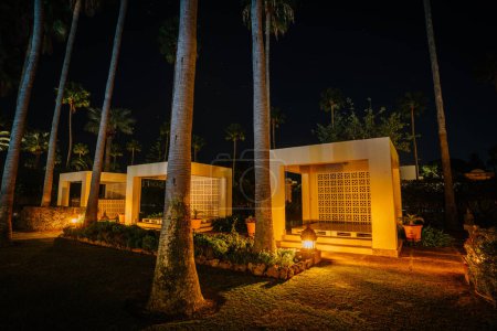 Sotogrante, Spain - January 27, 2024 - Night scene of a garden with tall palm trees and a lit gazebo-like structure with decorative wall patterns.