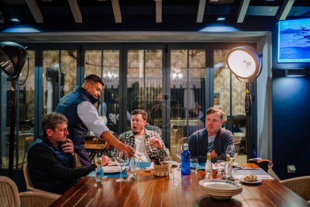 Foto de Sotogrante, España - 27 de enero de 2024 - Un camarero que sirve vino a los hombres en una mesa de restaurante, en el interior, noche, atuendo informal, comedor, conversación, bebidas, iluminación ambiental. - Imagen libre de derechos