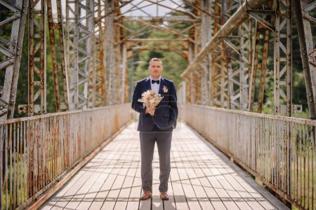 Valmiera, Latvia - July 7, 2023 - A man in a navy suit stands on a rusty metal bridge holding a bouquet, looking towards the camera.