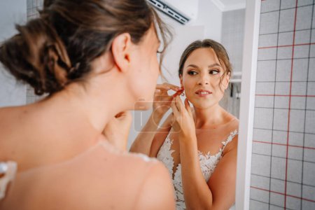 Téléchargez les photos : Valmiera, Lettonie - 7 juillet 2023 - Une mariée en robe de dentelle met sa boucle d'oreille en regardant son reflet dans un miroir. - en image libre de droit