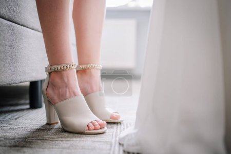 Valmiera, Latvia - July 7, 2023 - Closeup of a womans feet in open-toed beige high heels, with chunky gold ankle bracelets, standing by a chair and a white gown.