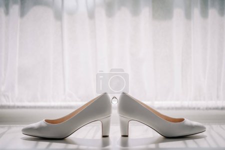 Foto de Valmiera, Letonia - 7 de julio de 2023 - Dos zapatos de novia blancos se colocan con un anillo de bodas equilibrado entre ellos contra una ventana con cortinas transparentes. - Imagen libre de derechos