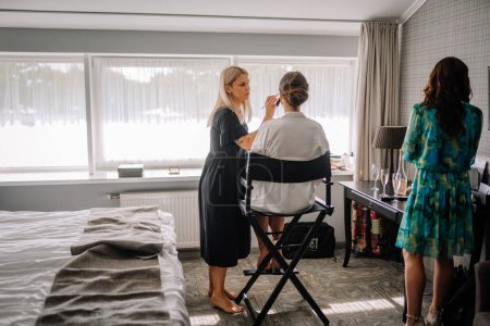 Valmiera, Latvia - July 7, 2023 - A makeup artist is working on a client in a director's chair in a room with another woman and champagne glasses.