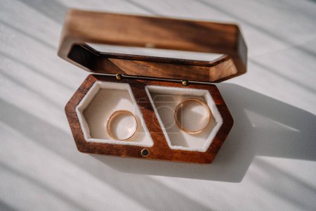 Valmiera, Latvia - July 7, 2023 - Two wedding rings in a hexagonal wooden box with white lining, on a white surface with shadows.