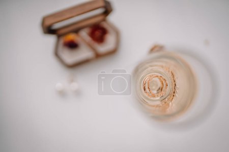 Foto de Valmiera, Letonia - 7 de julio de 2023 - Vista aérea de una copa de champán con anillos de boda en el interior, caja de anillos y pendientes en una superficie blanca. - Imagen libre de derechos