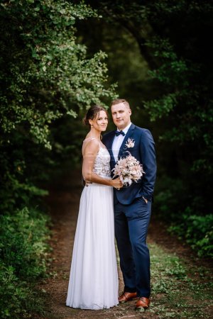 Valmiera, Latvia - July 7, 2023 - A bride and groom embrace in a forest, she in a white lace dress holding a bouquet, he in a blue suit and bow tie.