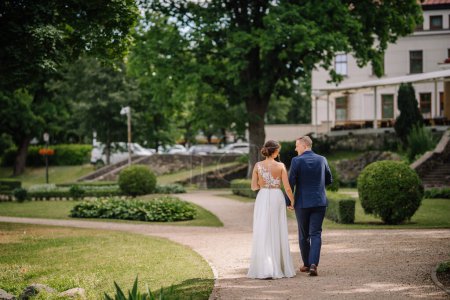 Valmiera, Lettland - 7. Juli 2023 - Braut und Bräutigam gehen Hand in Hand auf einem Gartenweg, mit viel Grün und einem Gebäude im Hintergrund.