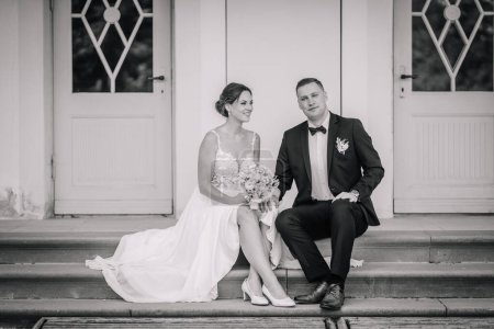 Valmiera, Latvia - July 7, 2023 - Bride and groom seated on steps, monochrome image, groom looks at camera, bride gazes at groom.