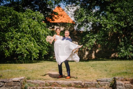 Valmiera, Lettland - 7. Juli 2023 - Ein Bräutigam hebt eine lächelnde Braut, während sie einen Strauß hält und ihr Kleid flattert; Grün und eine Backsteinmauer im Hintergrund.
