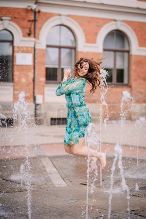 Photo for Valmiera, Latvia - July 7, 2023 - A joyful woman in a blue-green dress is captured mid-jump amidst water jets from a fountain, with a historic brick building in the background - Royalty Free Image