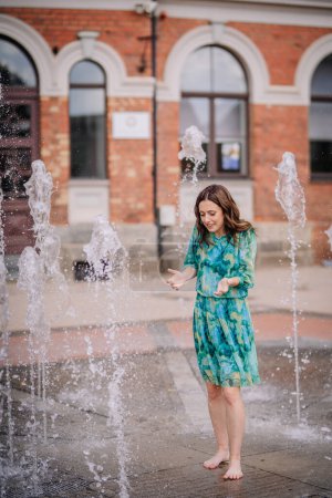 Téléchargez les photos : Valmiera, Lettonie - 7 juillet 2023 - Une femme en robe bleue et verte joue dans une fontaine, pieds nus, avec un bâtiment en brique et des fenêtres cintrées en arrière-plan. - en image libre de droit