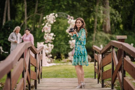Valmiera, Latvia - July 7, 2023 - A woman in a floral dress holds a camera on a wooden bridge, two people in the background.