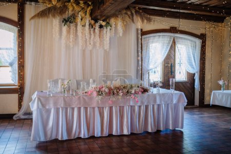 Valmiera, Latvia - July 7, 2023 - Wedding head table with floral centerpiece, candles, and hanging lights inside a rustic venue.