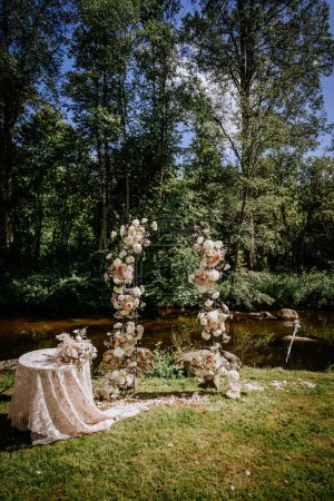 Valmiera, Lettland - 7. Juli 2023 - Floraler Hochzeitsbogen und Tisch an einem Fluss mit Waldkulisse.