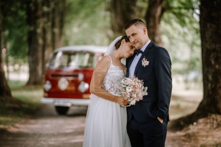 Valmiera, Latvia - July 7, 2023 - Bride and groom embracing in front of a vintage van in a wooded area.