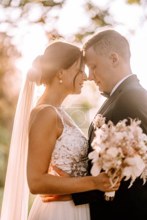 Valmiera, Latvia - July 7, 2023 - Bride and groom with foreheads touching, holding a bouquet, backlit by sunlight.