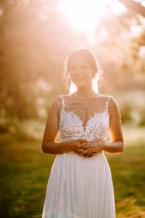 Foto de Valmiera, Letonia - 7 de julio de 2023 - Una novia radiante con un vestido de encaje blanco se encuentra en un parque iluminado por el sol, con las manos cerradas. - Imagen libre de derechos