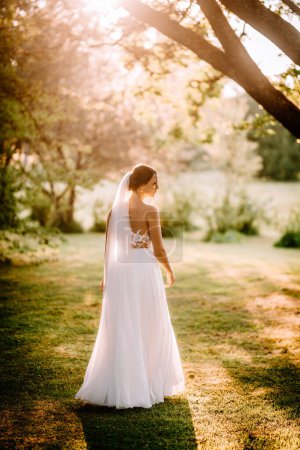 Valmiera, Latvia - July 7, 2023 - A bride in a white wedding dress and veil stands in a sunlit garden, looking away from the camera.