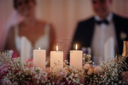Valmiera, Latvia - July 7, 2023 - Wedding table with candles in focus, floral arrangements, blurred couple in the background.