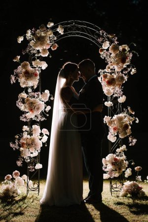 Valmiera, Lettonie - 7 juillet 2023 - Un couple silhouetté embrassant sous une arche florale la nuit, rétro-éclairé créant un effet dramatique.