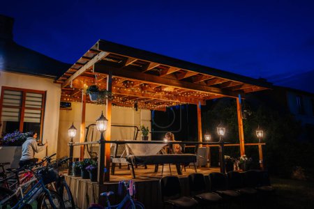 Photo for Valmiera, Riga - July 5, 2023 - Outdoor evening setting at a venue with string lights, wooden pergola, bicycles, and a person sitting at a table. - Royalty Free Image