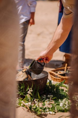 Jurmala, Latvia - july 25, 2023 - A person pours liquid from a black cup into a bowl on a bed of greenery and white blossoms, part of a beach ritual with folk elements visible.