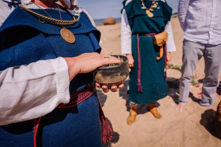 Jurmala, Latvia - july 25, 2023 - Close-up of a hand holding a small bowl with green leaves, part of a traditional ceremony, with people in folk costumes in the background on a sandy beach.
