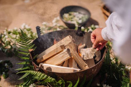 Jurmala, Latvia - july 25, 2023 - A hand is lighting a fire in a rustic cauldron surrounded by green ferns and white flowers on sandy ground.