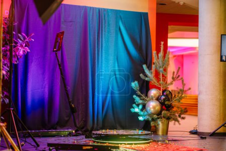 Sigulda, Lettland - 12. Januar 2024 - Leerer Bühnenbereich mit blauem Vorhang, LED-Lampen, Mikrofonständer, kleinem Weihnachtsbaum mit Ornamenten und Konfetti auf dem Boden.