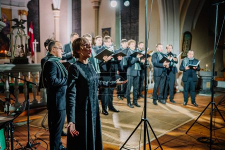 Téléchargez les photos : Valmiera, Lettonie - 7 janvier 2024 - Église Saint-Simon. Une femme chantant en solo avec une chorale en arrière-plan à l'intérieur d'une église, des micros et de la musique est visible. - en image libre de droit
