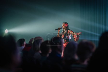 Foto de Valmiera, Letonia - 15 de febrero de 2024 - Una cantante femenina en un concierto tradicional gitano se presenta con un micrófono, resaltado por un foco de atención sobre un escenario malhumorado. - Imagen libre de derechos