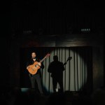 Valmiera, Latvia- February 15, 2024 - A male guitarist stands spotlighted on stage, casting a large shadow behind him, at a dimly lit gypsy traditional concert.