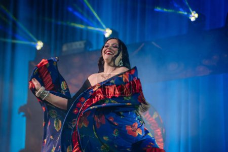 Foto de Valmiera, Letonia - 15 de febrero de 2024 - Una bailarina alegre en un colorido disfraz gitano baila bajo luces azules en un concierto. - Imagen libre de derechos